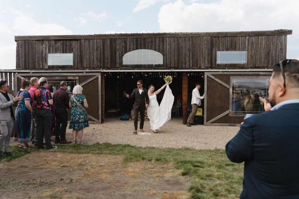 A couple step out through the big barn doors as guests look on, Blue Sky Ranch wedding