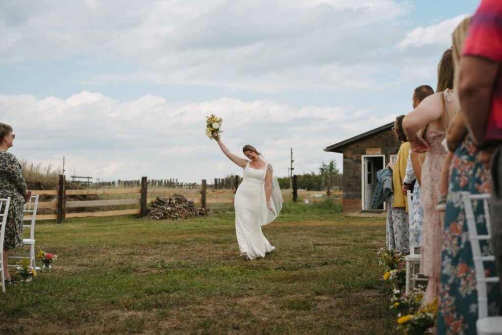 The bride walks across the yard toward the ceremony, bouquet raised, Blue Sky Ranch wedding