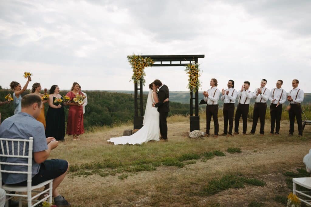 A couple kiss under a timber arbour on the ravine edge with the wedding party beside them, Blue Sky Ranch wedding